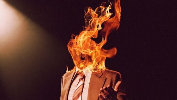 Cropped image of a man in a brown suit and tie whose face is engulfed in fire from the Late Night With The Devil official movie poster.